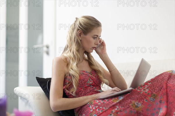 Woman in dress sitting on sofa and using phone and laptop