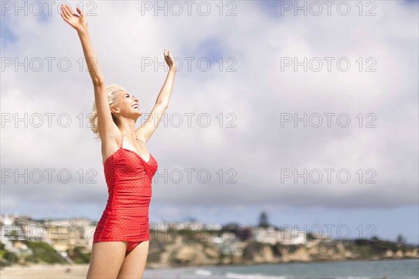 Woman wearing red one piece swimsuit standing on beach with arms raised