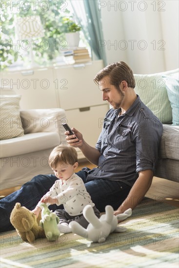 Father sitting on floor with little son (2-3 years) in living room and texting message.