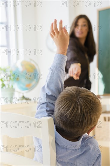 Female teacher standing beside blackboard in classroom, schoolboy (6-7) with hand raised