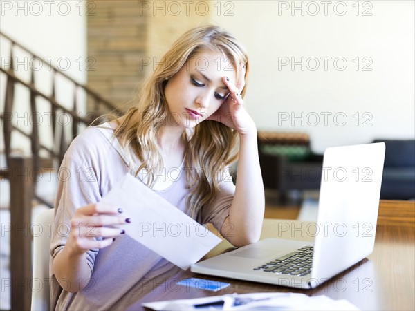 Young woman sitting at table with laptop looking at paper bill