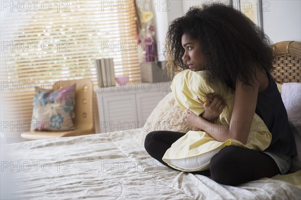 Teenage girl (14-15) sitting on bed, embracing pillow.