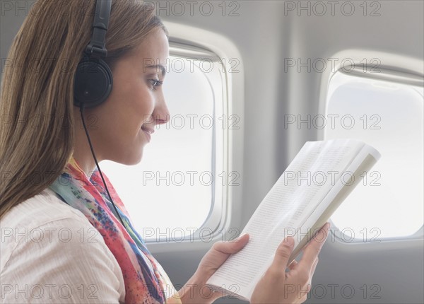 Young woman wearing headphones while reading book on plane.