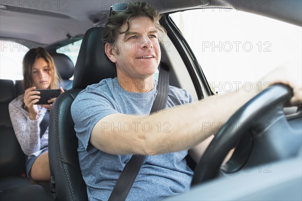 Man driving car, girl (10-11) texting in background.