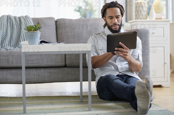 Young man with dreadlocks using digital table by sofa in living room.