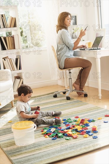 Boy (2-3) playing with toy blocks while mother working on laptop.