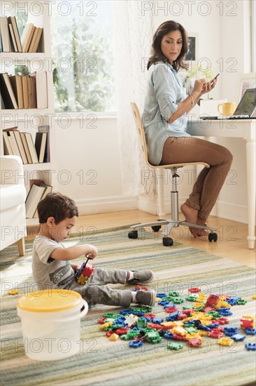 Boy (2-3) playing with toy blocks while mother working on laptop.