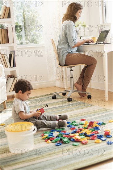 Boy (2-3) playing with toy blocks while mother working on laptop.