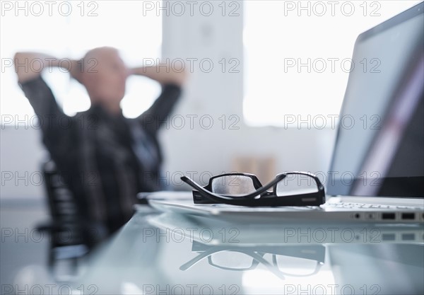 Eyeglasses and laptop on desk in office, man relaxing in background.