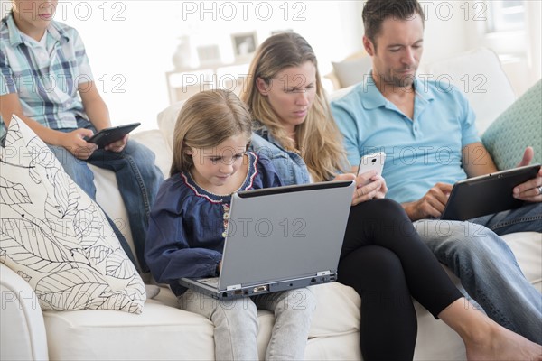 Family with two children (6-7, 8-9) sitting on sofa, using laptop and digital tablets