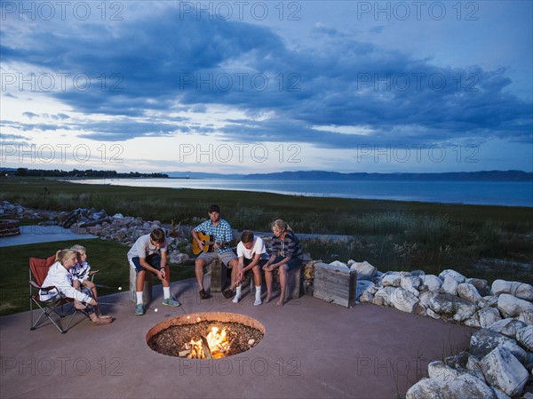 Father with children (8-9, 10-11, 16-17) sitting in front of campfire and playing guitar