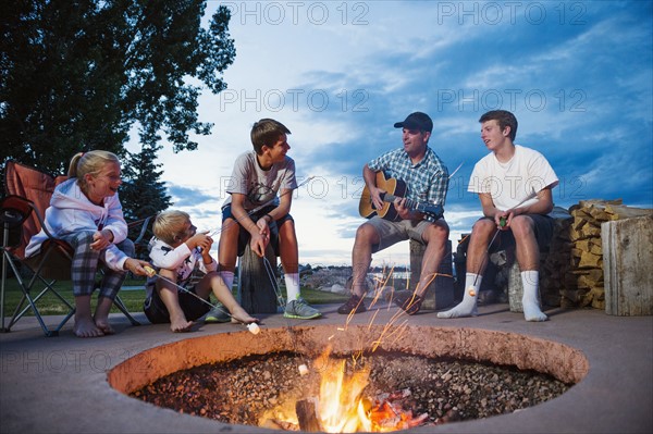 Father with children (8-9, 10-11, 16-17) sitting in front of campfire and playing guitar