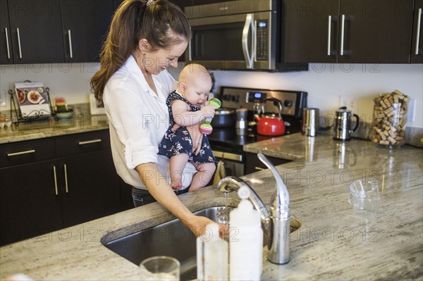 Mother holding baby daughter (2-5 months) and filling glass with water.