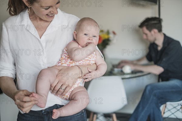 Mother holding baby (2-5 months) and father working in background.