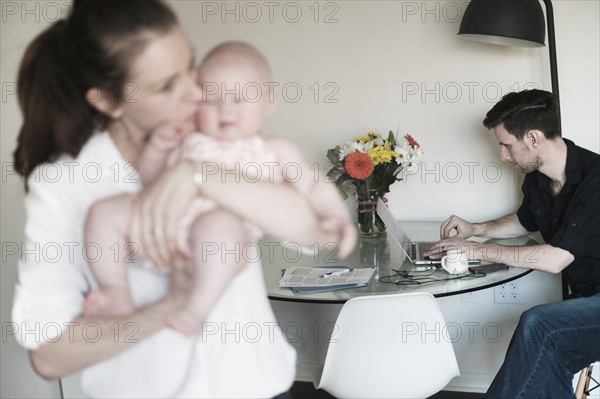 Mother holding baby (2-5 months) and father working in background.