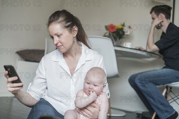Mother holding baby (2-5 months) while using smart phone and father working in background.