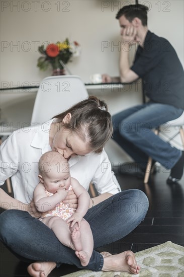 Mother holding baby (2-5 months) and father working in background.