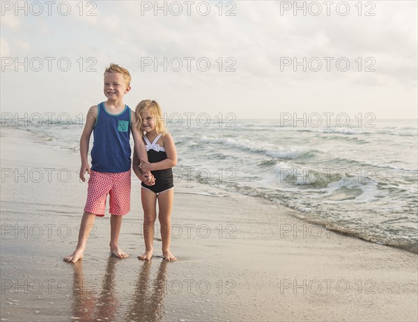 Boy (6-7) and girl (4-5) standing on beach by water holding hands