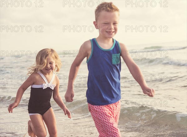 Boy (6-7) and girl (4-5) running on beach by water