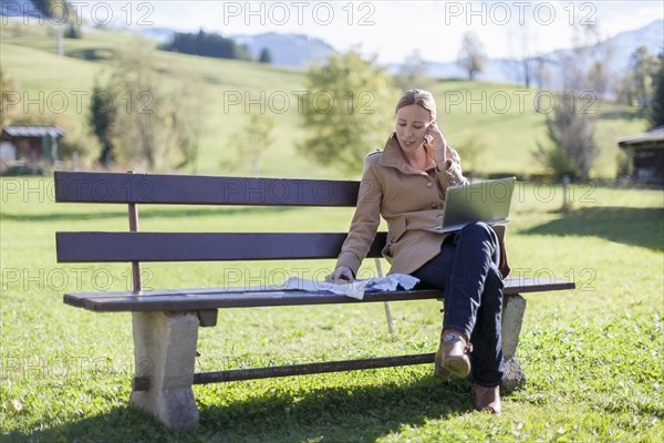 Austria, Salzburger Land, Maria Alm, Mature woman sitting on bench and talking on phone