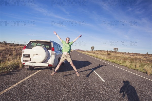 Australia, Outback, Northern Territory, Red Centre, Uluru-Kata Tjuta National Park, Woman jumping next to SUV car on road in wilderness