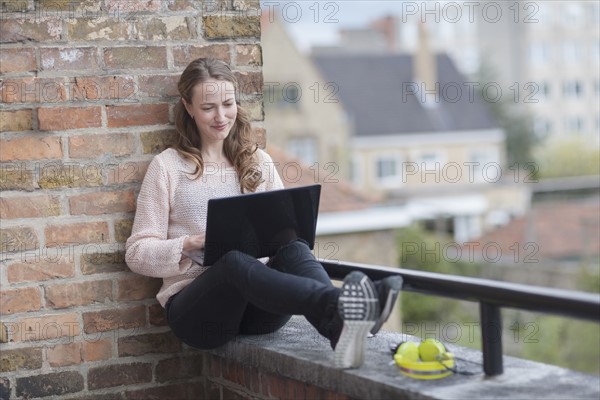 Mid-adult woman sitting on balcony railing and using laptop