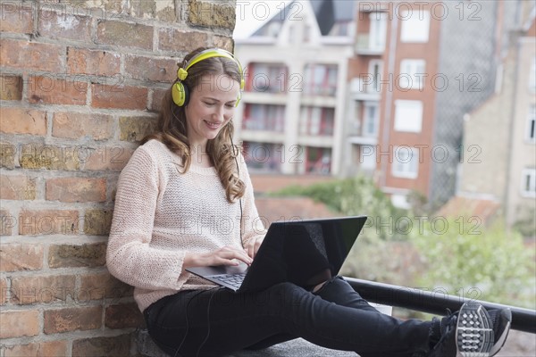Mid-adult woman with headphones on sitting on balcony railing and using laptop