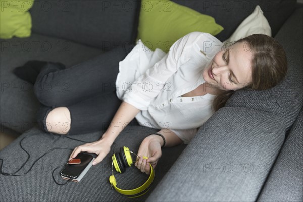Mid-adult woman lying on sofa and holding smart phone and headphones