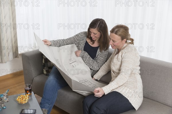 Smiling women sitting on sofa in living room and looking at blueprint