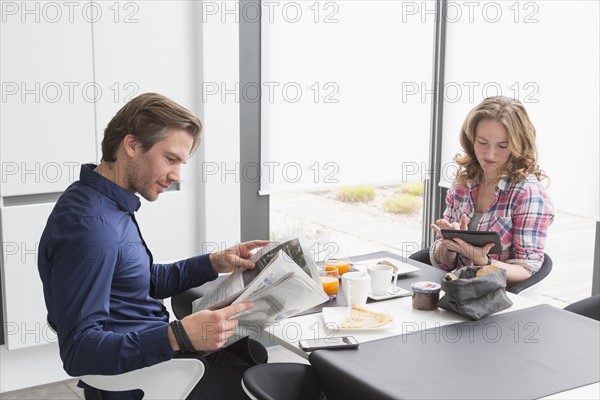 Mid-adult couple reading newspaper and using tablet at breakfast table