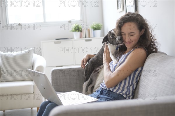 Young woman with laptop cuddling with her dog on sofa.