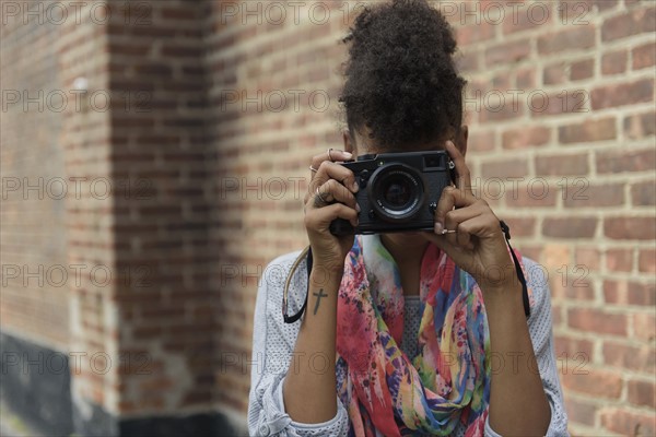 Young woman taking picture with digital camera against brick wall.