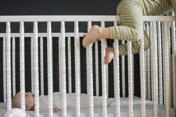 Girl (2-3) climbing on crib and brother (6-11 months) lying inside