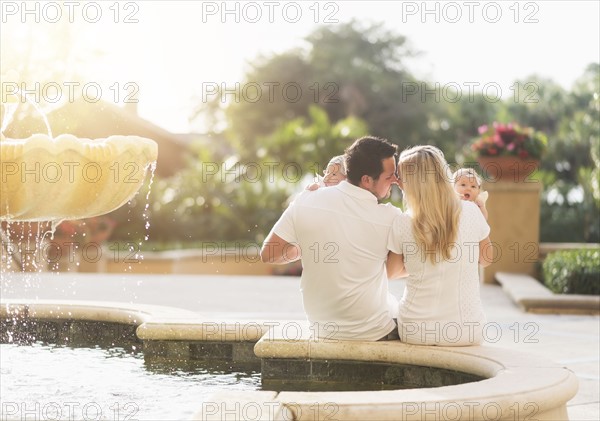 Happy family with two baby girls (2-5 months) near fountain in sunlight
