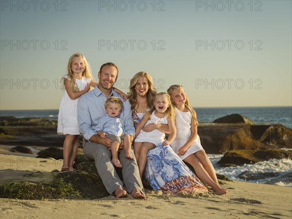 Family with children (12-17 months, 4-5, 6-7, 8-9) sitting on beach with sea in background