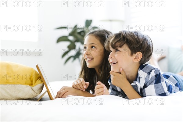 Siblings (10-11, 6-7) lying in bed and using tablet