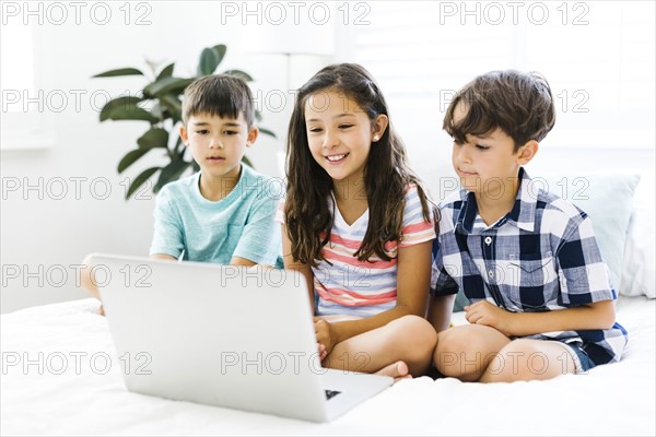 Siblings (10-11, 6-7, 8-9) sitting in bed and using computer