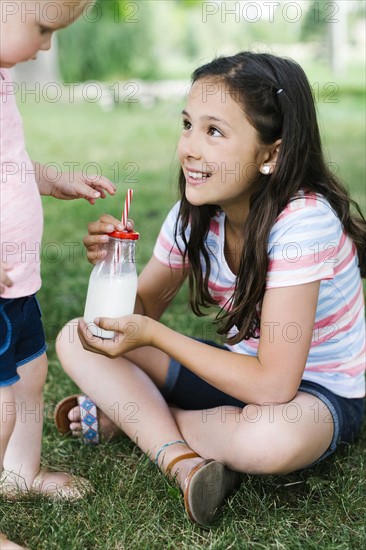 Girl (10-11) sharing bottle of milk with sister (2-3) in park