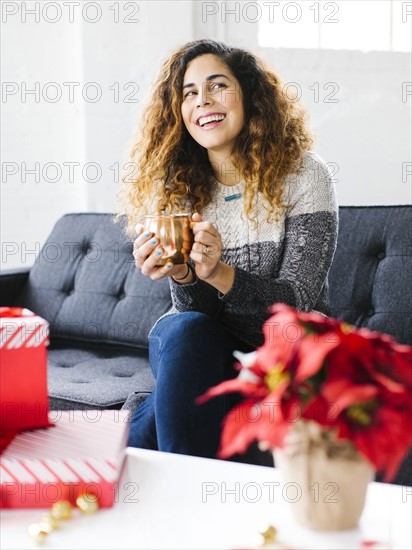 Happy woman on sofa, holding mug with Christmas gifts in foreground