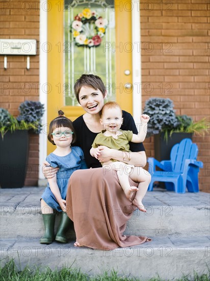 Mother with daughters (2-3, 12-17 months) sitting in front of house