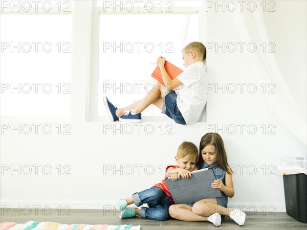 Boy (6-7) reading book and sister with brother (2-3, 6-7) using digital tablet