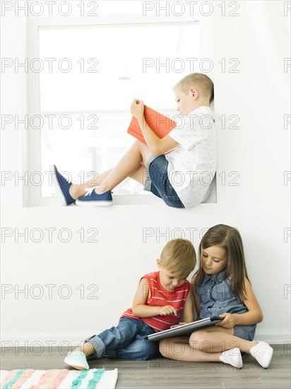 Boy (6-7) reading book and sister with brother (2-3, 6-7) using digital tablet