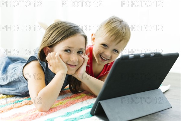 Portrait of siblings (2-3, 6-7) looking at camera and lying down on carpet next to digital tablet