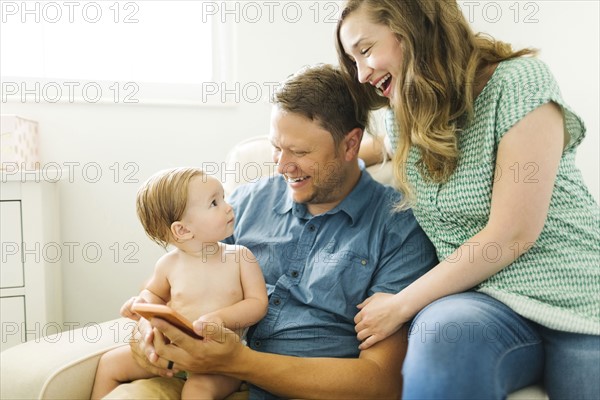 Mother and father with baby girl (12-17 months) sitting in living room and holding smart phone