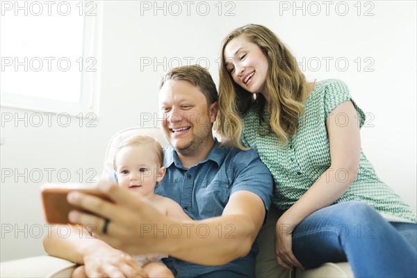 Mother and father with baby girl (12-17 months) sitting in living room and taking selfie