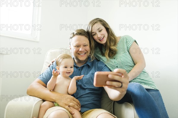 Mother and father with baby girl (12-17 months) sitting in living room and taking selfie