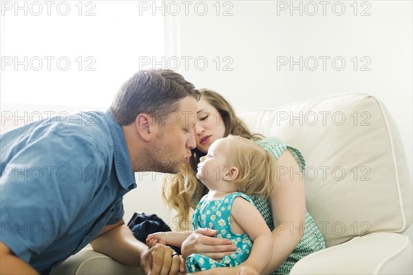 Mother and father with baby girl (12-17 months) sitting in living room and kissing