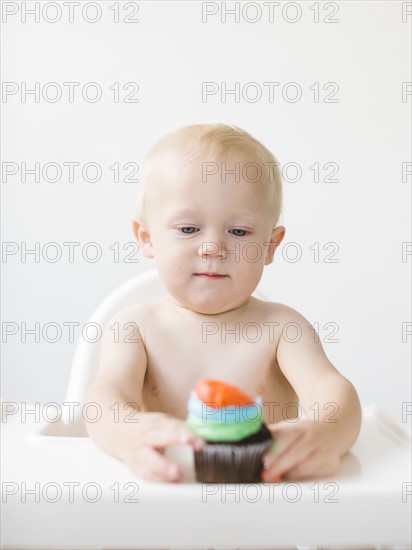 Baby boy (12-17 months) sitting in high chair and holding cupcake