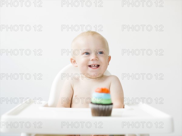 Baby boy (12-17 months) sitting in high chair with cupcake in front of him