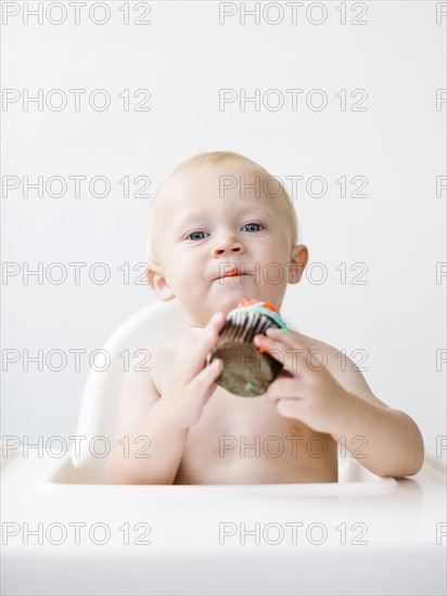 Baby boy (12-17 months) sitting in high chair and holding cupcake